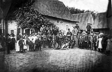 Bauernschützenfest Harsewinkel - Schützengesellschaft vor den Wohnhäusern des Schumachers Hermann Westhowe (im Hintergrund) und des Buchbinders und Fotografen Johann Hermann Jäger (rechts). Undatiert, um 1900?