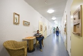 Auf dem Bild ist der Flur einer gesicherten Station zu sehen. An beiden Seiten sind Holztüren, die zu den Patientenzimmern führen. Im Vordergrund sieht man einen Tisch mit zwei Sesseln, in denen ein Patient sitzt. Auf dem Bild ist der Flur einer gesicherten Station zu sehen. An beiden Seiten sind Holztüren, die zu den Patientenzimmern führen. Im Vordergrund sieht man einen Tisch mit zwei Sesseln, in denen ein Patient sitzt.