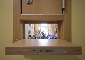 Auf dem Bild sieht man die offene Holzklappe eines Patientenzimmers, durch die man einen Patienten sieht, der auf einer Schreibmaschine schreibt Auf dem Bild sieht man die offene Holzklappe eines Patientenzimmers, durch die man einen Patienten sieht, der auf einer Schreibmaschine schreibt