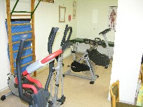 Auf dem Bild ist das Inneren eines Therapieraumes mit einigen Gerätschaften zu sehen. Auf einem der Geräte sitzt eine Physiotherapeutin. Auf dem Bild ist das Inneren eines Therapieraumes mit einigen Gerätschaften zu sehen. Auf einem der Geräte sitzt eine Physiotherapeutin.