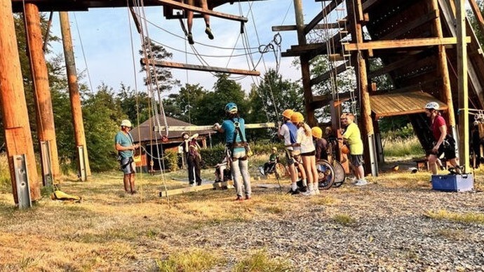 mehrere Erwachsene und Kinder mit Helmen stehen zwischen Holzpfosten und herabhängenden Seilen in einem Hochseilgarten mehrere Erwachsene und Kinder mit Helmen stehen zwischen Holzpfosten und herabhängenden Seilen in einem Hochseilgarten