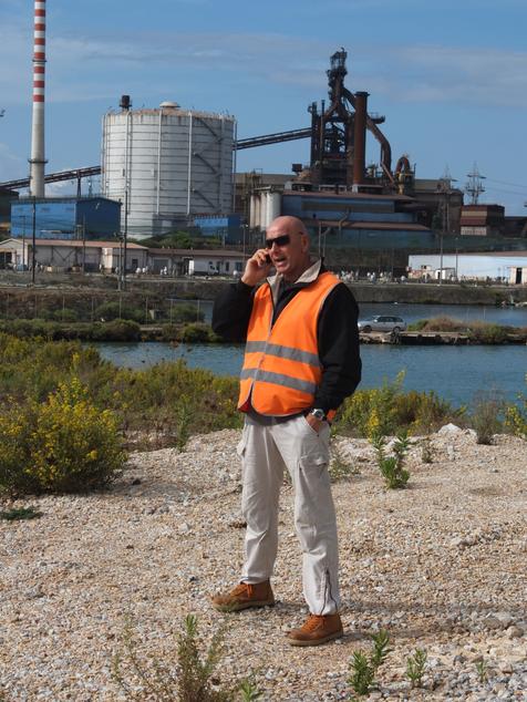 Ein Mann in orangener Sicherheitsweste steht vor einem Stahlwerk und telefoniert mit seinem Handy. Foto: Robert Laube (vergrößerte Bildansicht wird geöffnet)