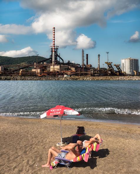Ein Mann und eine Frau liegen in ihren Sonnenstühlen am Strand, nur einen Steinwurf entfernt auf der anderen Uferseite steht ein Stahl- und Eisenwerk. Montage: Sabine Korth (vergrößerte Bildansicht wird geöffnet)