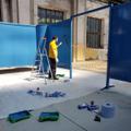 Ein Mann in schwarzer Hose und gelben Hemd streicht eine Aufstellwand in blau. (vergrößerte Bildansicht wird geöffnet)