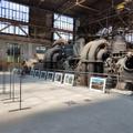 Gerahmte Fotos lehnen gegen die Absperrung einer stillgelegten Industriemaschine, während davor freistehende, leere Ständer für die Bilder zu erkennen sind. (vergrößerte Bildansicht wird geöffnet)