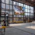 An der Fensterfront einer Industriehalle hängt ein großformatiges Foto mit einem Naturmotiv, unter der Fensterfront stehen gerahmte Bilder, die noch nicht an ihrem Platz angebracht wurden und links steht eine Frau an zwei behelfsmäßig provisorisch aufgestellten Tischen. (vergrößerte Bildansicht wird geöffnet)