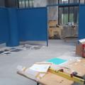 Auf einem Tisch liegen ein geöffneter Karton, eine Wasserwaage und Verpackungsmaterial während im Hintergrund eine blaue Stellwand zu sehen ist, um die herum Bilder liegen. (vergrößerte Bildansicht wird geöffnet)