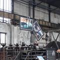 Zwei auf schwarzen Pappen gedruckte Collagen hängen von der Decke einer Industriehalle, von denen die eine bisher nur an einer Kette festgemacht wurde und deshalb schiefhängt. (vergrößerte Bildansicht wird geöffnet)