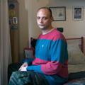 Ein Mann sitzt in einem spärlich eingerichtetem Zimmer auf seinem Bett. (vergrößerte Bildansicht wird geöffnet)