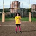Eine junge Frau in Sportbekleidung steht vor einem Rugbytor, dahinter liegen Hochhäuser. (vergrößerte Bildansicht wird geöffnet)