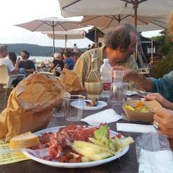 Eine Gruppe von Menschen sitzt an einem Tisch am Strand und isst eine kleine Mahlzeit. (vergrößerte Bildansicht wird geöffnet)