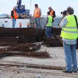 Ein Mann richtet seine Kamera auf eine Gruppe von Arbeitern, die auf einer Baustelle am Meer herumstehen. (vergrößerte Bildansicht wird geöffnet)