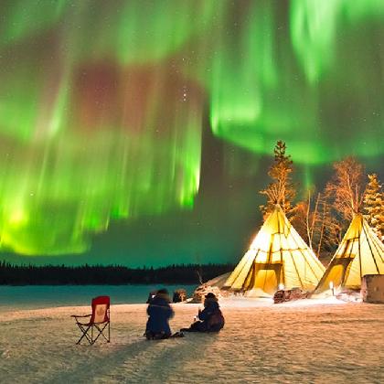 Menschen am Feuer im Schnee vor Zelten sitzend, über ihnen leuchten Polarlichter.