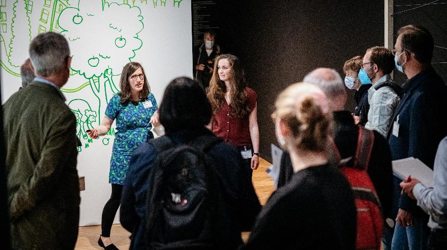 Menschen stehen zusammen und hören einer Frau zu, die was erklärt.