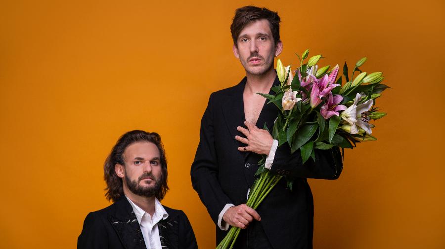 Bild der Künstler. Die beiden sind in schwarz gekleidet, einer hält Blumen im Arm.