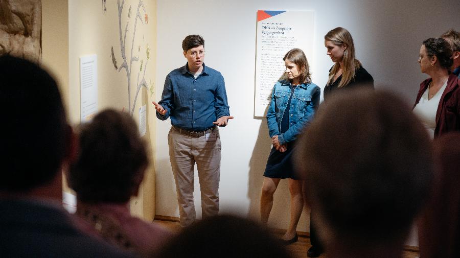 Eine Frau steht vor einer Infotafel im Museum und erklärt einer Personengruppe etwas.