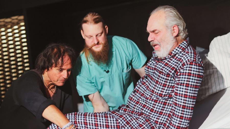 Zwei Männer helfen einem älteren Mann im karierten Schlafanzug beim Aufsetzen auf einem Bett.
