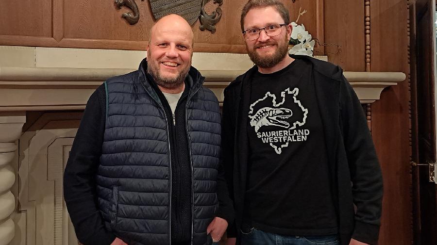 Stefan Schröder und Achim Schwermann stehen nebeneinander vor einer holzvertäfelten Wand. 