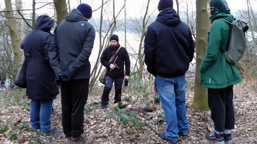Gruppe von Menschen steht im Wald und hört einer Person zu, die etwas erklärt.