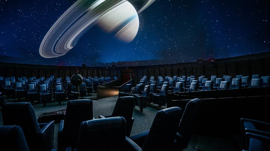 Der Planetariumssaal mit Sesseln und dem Projektor. Die Kuppel des Planetariums zeigt den Saturn