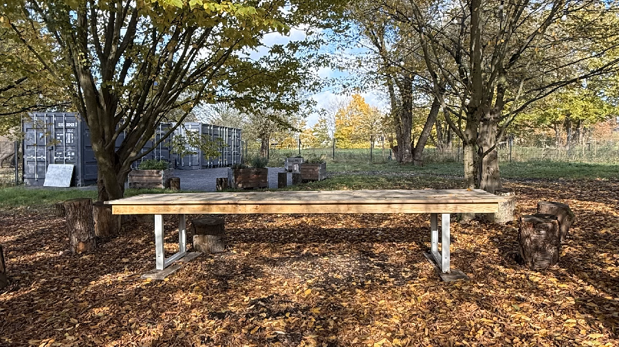 Auschnitt des Museumsgartens: Großer Holztisch auf laubbedecktem Boden unter zwei Bäumen