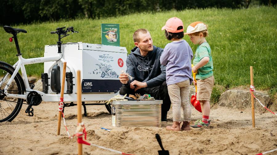 Ein Erwachsener zeigt zwei Kindern auf einem Spielplatz einen Knochen. Daneben steht ein Lastenrad.