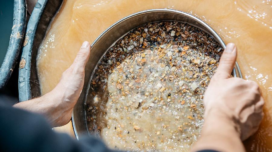 Person hält ein Metallsieb mit Kies und Sediment im trüben Wasser und siebt das Material aus.