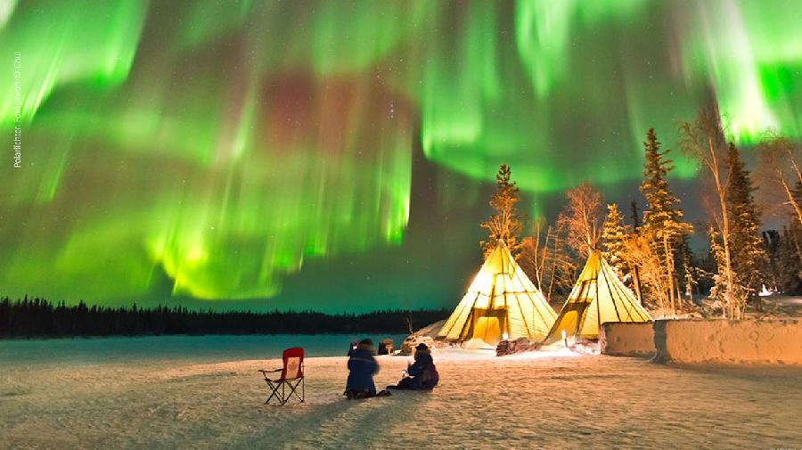 Menschen am Feuer im Schnee vor Zelten sitzend, über ihnen leuchten Polarlichter.