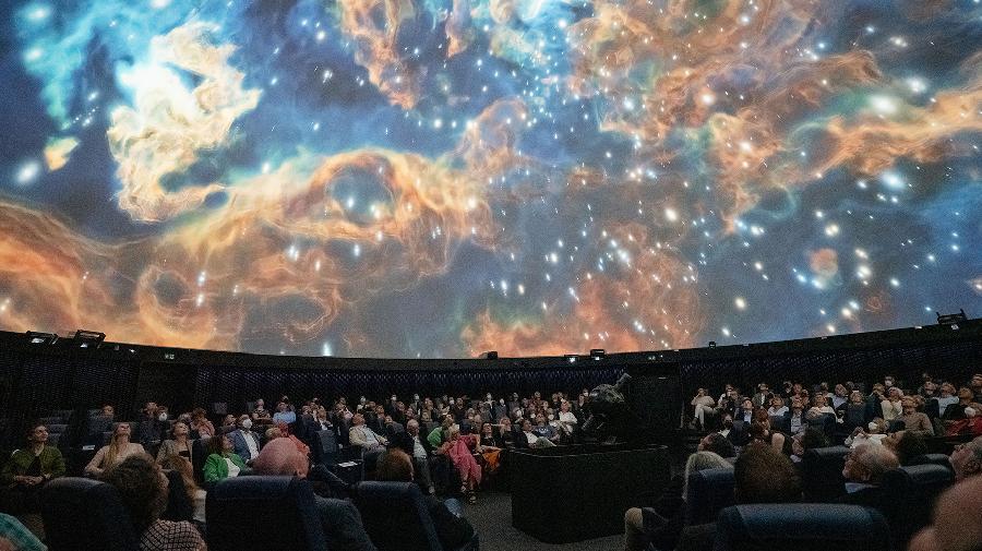 DIe Kuppel des Planetariums zeigt einen Sternennebel. Darunter sitzt das Publukum.