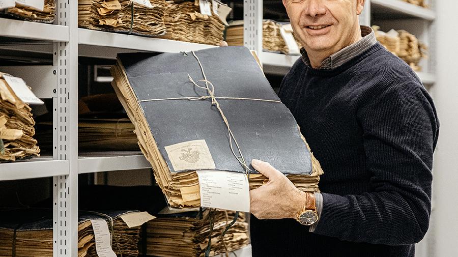 Mann hält einen großen historischen Aktenbestand in einem Archiv, umgeben von Regalen.