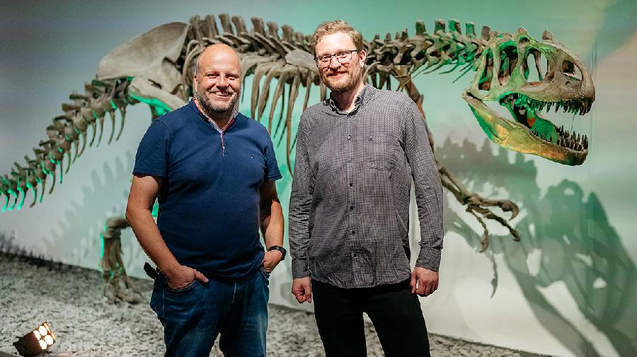 Zwei Männer stehen vor einem Dinoskelett und lächeln in die Kamera.