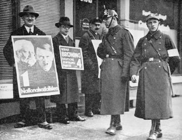 Wahlplakate vor einem Berliner Wahllokal vor der Reichstagswahl am 5. März 1933. Im Vordergrund ein Schutzpolizist und ein als Hilfspolizist gekennzeichneter SA-Mann.

Fotograf: A. Groß