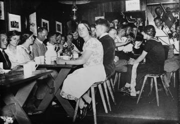 Richard Schirrmann (am zweiten Tisch links) mit musizierender Jugendgruppe im Tagesraum einer Jugendherberge (?), ohne Ort, undatiert, 1920er Jahre?