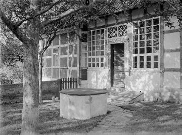 Sproßenfensterfassade eines Fachwerkhauses und Eingangstür mit Inschrift. Vorn: Brunnen und Kellerklappe. Vermutlich Raum Enger. Ohne Angaben, undatiert, 1940er Jahre?