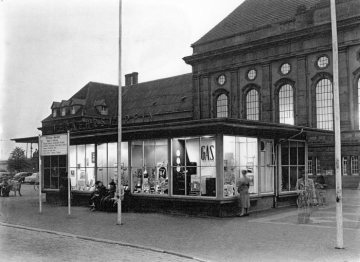 Bahnhof Hamm: Omnibus-Bahnhof und Gebäude des Verkehrsvereins mit Werbeschaufenstern örtlicher Geschäfte - darunter: Fotogeschäft (links) und Spirituosen (Mitte). Undatiert, um 1955 [?]