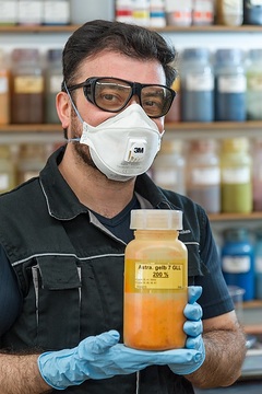 Schmitz Textiles GmbH & Co. KG: Im Labor – Porträt des Musa Atalay mit einer Farbpigment- Flasche „Astra Gelb“ für die nächste Textilfärbung