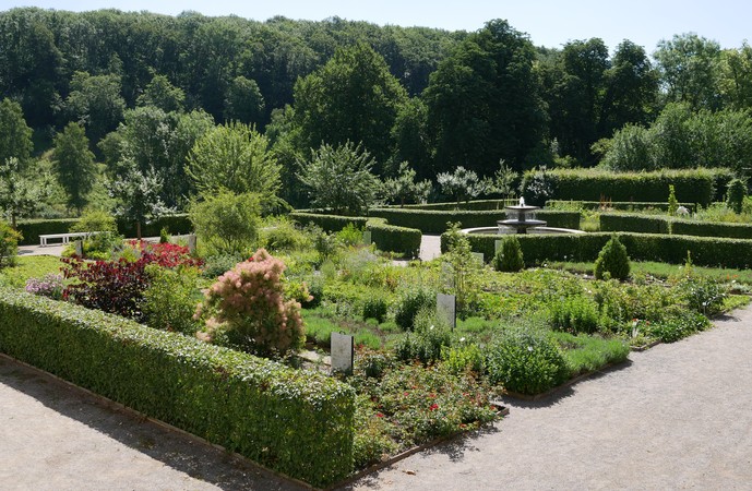 In einem Garten mit symmetrisch angelegten Beeten und Hecken befindet sich ein Stringbrunnen. Im Hintergrund befindet sich ein Laubwald.