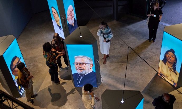 Blick ins Foyer der Ausstellung mit Bildschirmen, auf denen Porträts zu sehen sind. Blick ins Foyer der Ausstellung mit Bildschirmen, auf denen Porträts zu sehen sind.