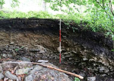 Das Tal der Brachtenbecke (Altena/ Märkischer Kreis) wurde über zwei Meter tief ausgespült und dabei ein Schlackenplatz teilzerstört. In der Böschung sind die dunklen Fundschichten dieser mittelalterlichen Verhüttungswerkstatt und einer darüber liegenden Meilerschicht zu erkennen.&lt;br&gt;Foto:LWL/D. Riemenschneider 