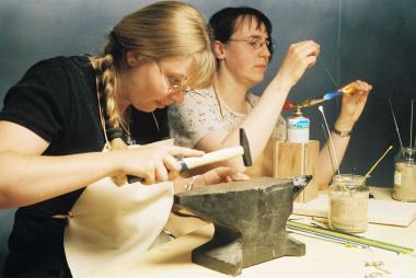 Alexandra Boßmann (vorne) und Marianne Gorissen vom Team der Museumspädagogen fertigen silberne Gewandspangen auf einem Amboss und schmelzen Glasperlen.<br>Foto. LWL/Brentführer.</br> 