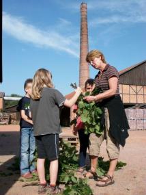 Museumspädagogin Charlotte Gaus hilft den Kindern beim Zuschneiden von Naturmaterialien vor dem Ringofen der Ziegelei. <br>Foto: LWL/Buba<br />