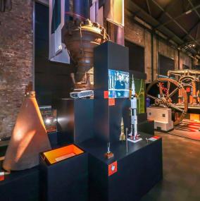 Blick in die Ausstellungsabteilung "Wissen wollen. Von Profit und Moral" mit Originalteilen von V2-Raketen sowie Modellen von US-Raketen aus den 1950er und 60er Jahren. Auf die Fahnen, die von der Decke herabhängen, laufen Filmsequenzen des Starts von Apollo 11.<br>Foto: LWL / Schütze