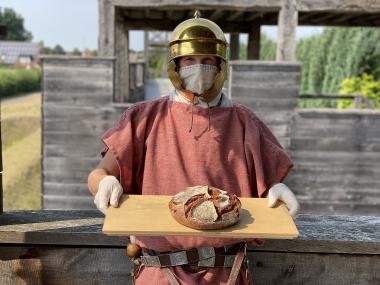 Ein behelmter Legionär mit Mundschutz und Handschuhen reicht frisch gebackenes Brot aus dem Feldbackofen.<br>Foto: LWL-Römermuseum/J. Mühlenbrock