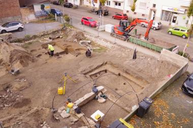 Blick auf die gesamte Grabungsfläche von Südosten. Die zwei größeren, eingetieften Grundrisse stellen die sich überschneidenden Grubenhäuser dar. Im Hintergrund mehrere runde gemauerte Brunnen. Im Vordergrund unter dem Eisengestell der gemauerte Rest eines Kellers aus behauenen Sandsteinen, die zu einem nicht mehr existierenden Barockgebäude zur Heierstraße hin gehören. <br>Foto: Denkmal3d/Dieter Lammers