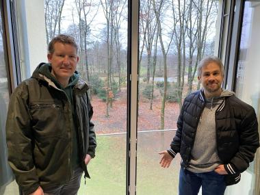 David Schulte-Döinghaus (l., Leiter der Gärtnerei des LWL-Klinikums Gütersloh) und Ulrich Paschke (Untere Denkmalbehörde der Stadt Gütersloh) im Gebäude 64 (Klinik für Psychosomatische Medizin und Psychotherapie) mit einigen der zu fällenden Bäumen im Hintergrund.<br>Foto: LWL/Dresmann